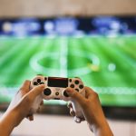 Close-up of hands holding a game controller playing a football video game on screen.